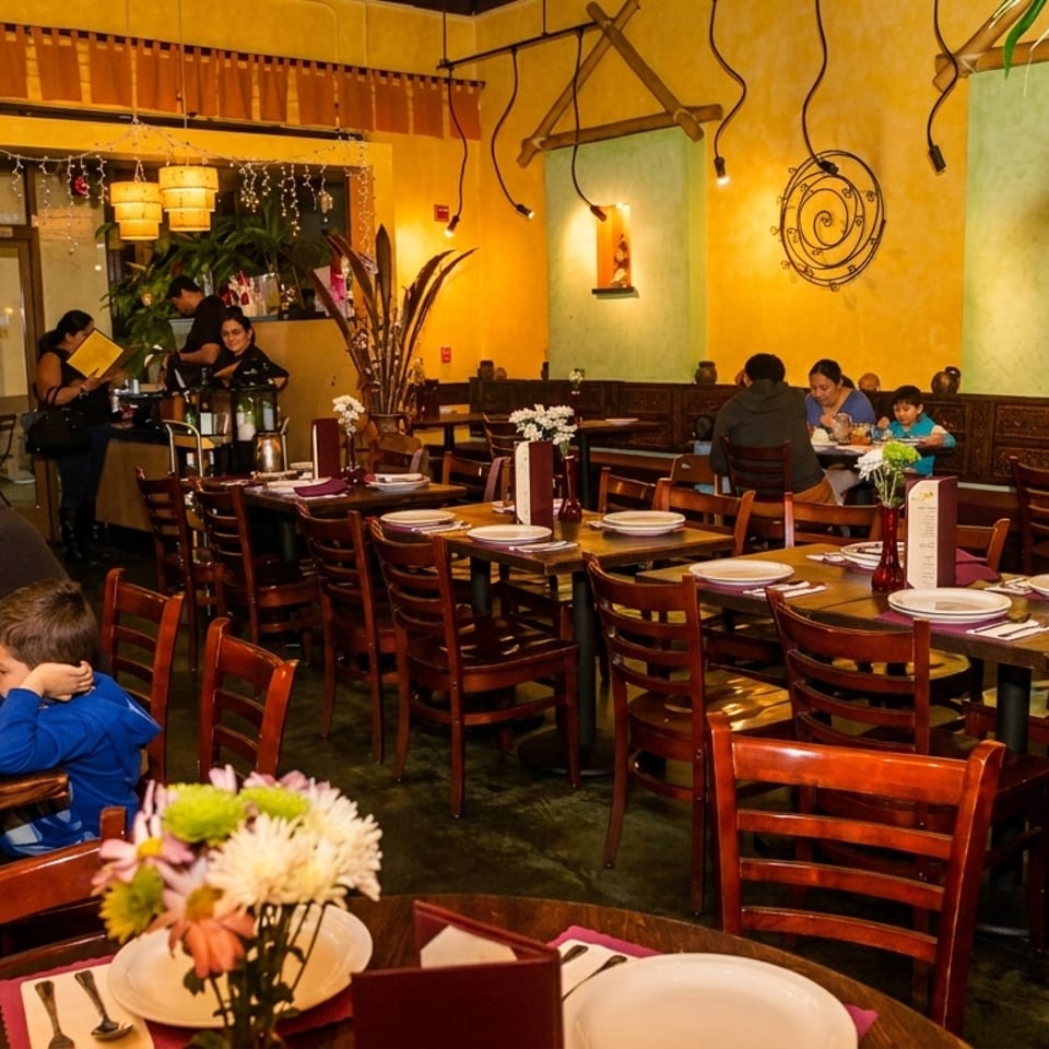Cozy restaurant interior with families dining at wooden tables, featuring warm yellow walls and bamboo plants.